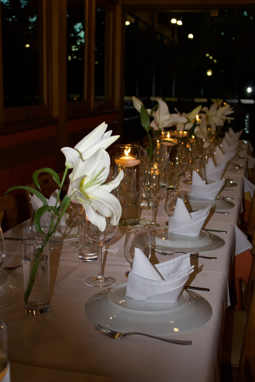 Lírios e velas na mesa longa — Mesa posta em branco absoluto com lírios altos, taças e velas flutuantes.